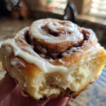 A hand holding a freshly baked, soft cinnamon bun generously topped with white cream cheese icing.