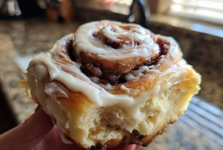 A hand holding a freshly baked, soft cinnamon bun generously topped with white cream cheese icing.