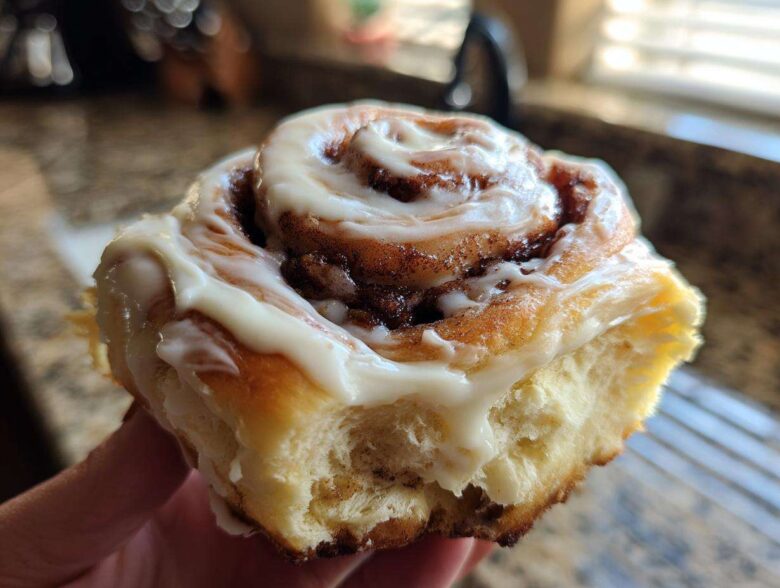 A hand holding a freshly baked, soft cinnamon bun generously topped with white cream cheese icing.