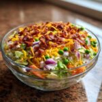 A clear glass bowl filled with Amazing 7 Layer Salad, topped generously with shredded cheddar, bacon bits, peas, and red onion.