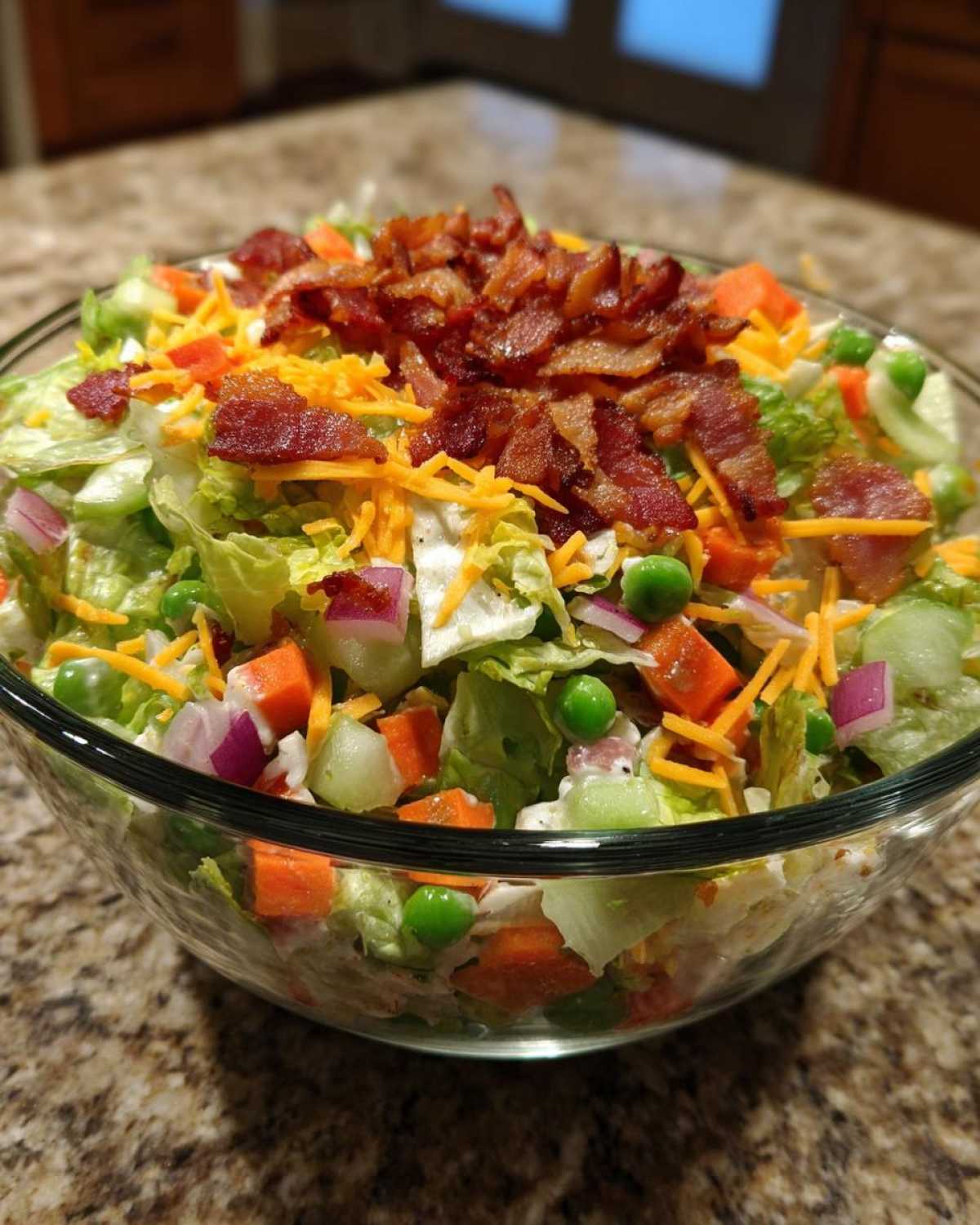 A close-up view of the top layer of an Amazing 7 Layer Salad featuring crisp lettuce, shredded cheese, peas, carrots, red onion, and crumbled bacon.
