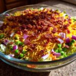 Close-up of a clear glass bowl filled with Amazing 7 Layer Salad, featuring layers of lettuce, peas, red onion, shredded cheese, and bacon bits.