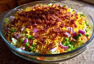 Close-up of a clear glass bowl filled with Amazing 7 Layer Salad, featuring layers of lettuce, peas, red onion, shredded cheese, and bacon bits.