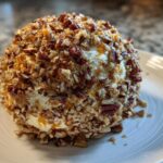 A perfectly formed, creamy cheese ball recipe coated entirely in chopped pecans, resting on a white plate.