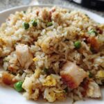A close-up shot of a serving of flavorful chicken fried rice with visible pieces of chicken, scrambled egg, and green peas.