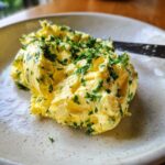 A dollop of creamy, yellow garlic butter mixed with chopped green herbs, served on a speckled white plate.