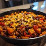Close-up of a hearty sweet potato skillet dinner with ground meat, black beans, and melted cheddar cheese in a black cast iron pan.