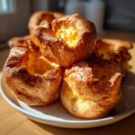 A close-up of several golden brown, perfectly risen yorkshire pudding pastries piled on a white plate.