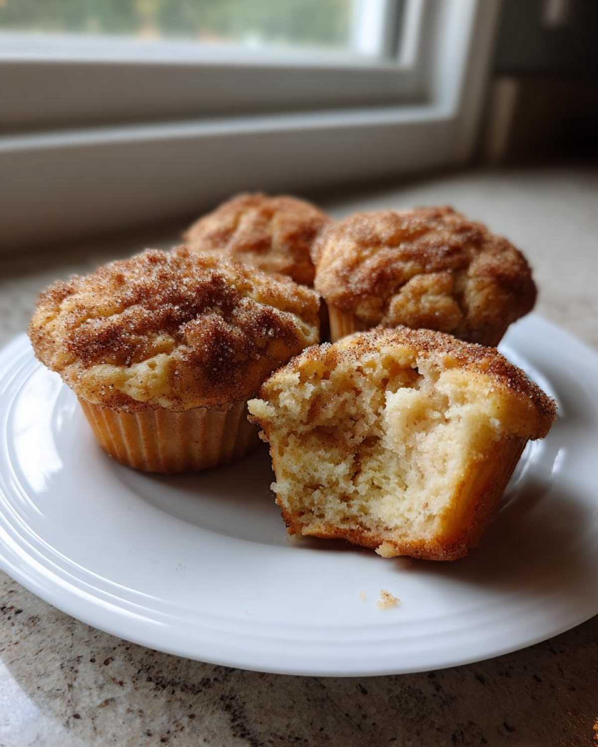 Four Apple Cinnamon Greek Yogurt Muffins on a white plate, one broken open showing the soft interior.