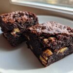 Two rich, fudgy Banana Bread Brownies with Walnuts stacked on a white plate near a window.
