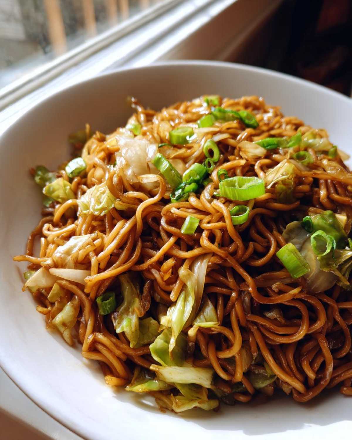 A close-up shot of a white bowl filled with glossy, brown fried noodles mixed with sautéed cabbage and topped with fresh green onions.