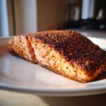 A close-up of a perfectly cooked piece of blackened salmon resting on a white plate, showing a dark spice crust.