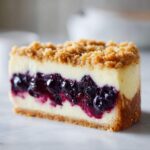 A perfect slice of Blueberry Crumble Cheesecake showing layers of graham cracker crust, creamy filling, blueberry swirl, and crumb topping.