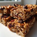 A stack of chewy homemade granola bars featuring oats, dried fruit, and chocolate chips, brightly lit.