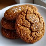 Close-up of several Chewy Salted Honey Cookies stacked on a white plate, topped with visible flaky sea salt crystals.