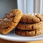 A stack of three chewy salted honey cookies, topped with flaky sea salt crystals, resting on a white plate.