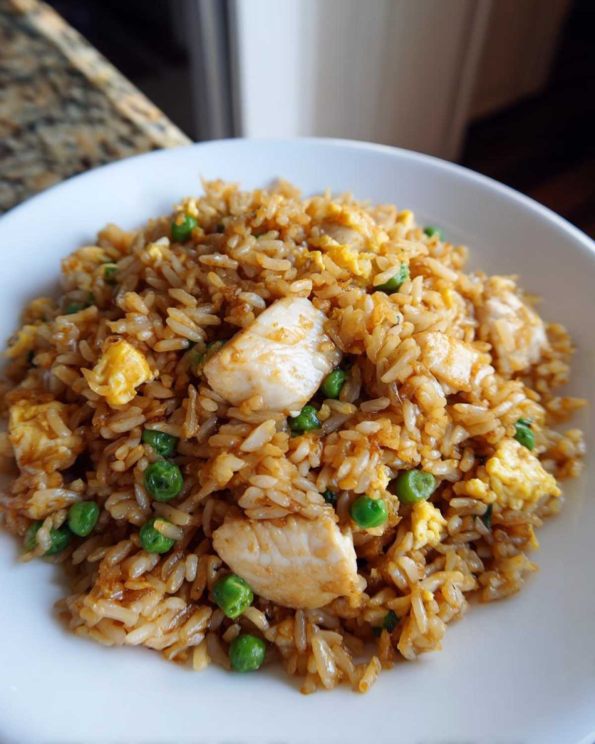 A close-up view of a white bowl filled with savory chicken fried rice, featuring chunks of chicken, scrambled egg, and green peas.