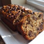 A golden brown loaf of The Chocolate Chip Banana Bread So Moist! with one slice cut and showing rich chocolate chips inside.
