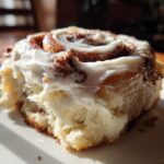 Close-up of a fluffy cinnamon roll generously topped with thick, white cream cheese frosting.