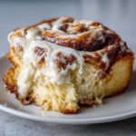 A close-up of a fluffy cinnamon roll generously topped with thick, dripping cream cheese frosting.