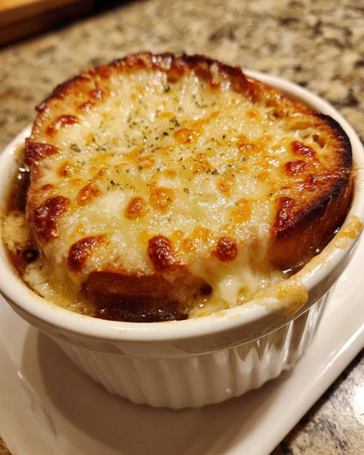 Close-up of a white crock filled with bubbling french onion soup topped with a toasted crouton and thick layer of melted, browned cheese.
