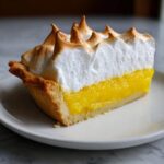 A perfect slice of classic lemon pie showing bright yellow filling and tall, toasted meringue topping.