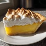 A perfect slice of classic lemon pie featuring bright yellow filling and tall, toasted meringue topping on a white plate.
