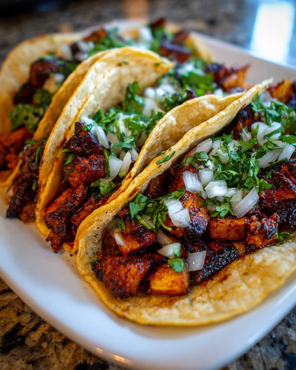 A close-up view of three street-style Tacos al pastor served on small corn tortillas, topped with marinated pork, white onion, and cilantro.