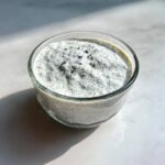 A close-up of creamy coconut chia pudding in a clear glass bowl, set on a bright white surface.