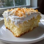 A close-up of a moist slice of coconut poke cake topped with whipped cream and toasted coconut flakes.