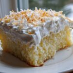 A moist slice of coconut poke cake topped with whipped cream and toasted coconut flakes.