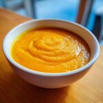 A close-up of a bowl filled with thick, vibrant orange Cozy Butternut Squash Soup, resting on a wooden surface.