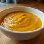 A close-up of a bowl filled with thick, vibrant orange Cozy Butternut Squash Soup resting on a wooden countertop.