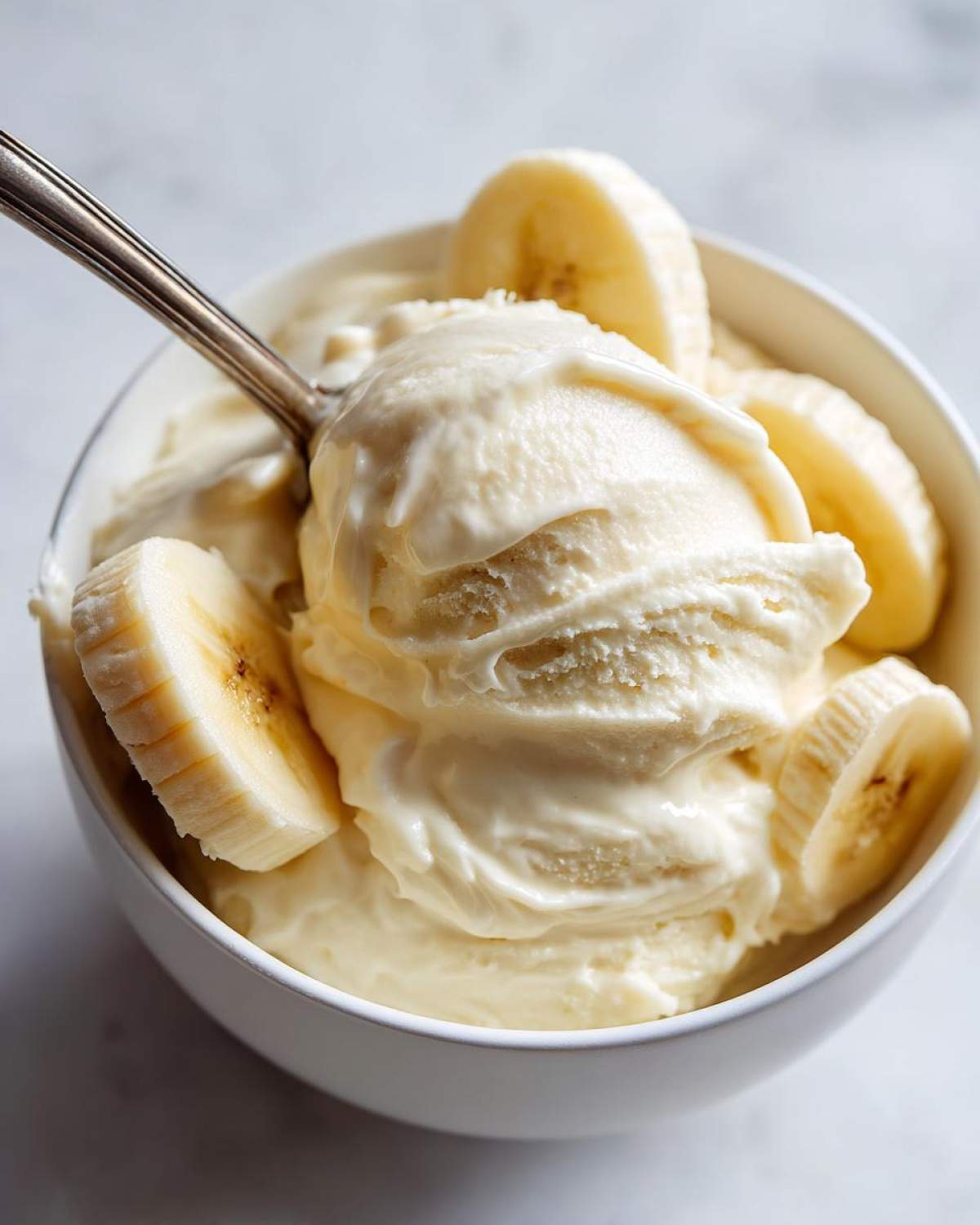 A close-up of creamy banana ice cream in a white bowl, topped with fresh banana slices, with a spoon scooping some out.