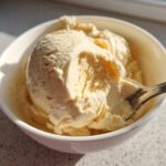 A close-up of creamy, pale yellow banana ice cream scooped into a white bowl with a spoon resting in it.