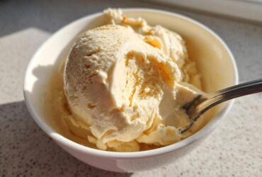 A close-up of creamy, pale yellow banana ice cream scooped into a white bowl with a spoon resting in it.