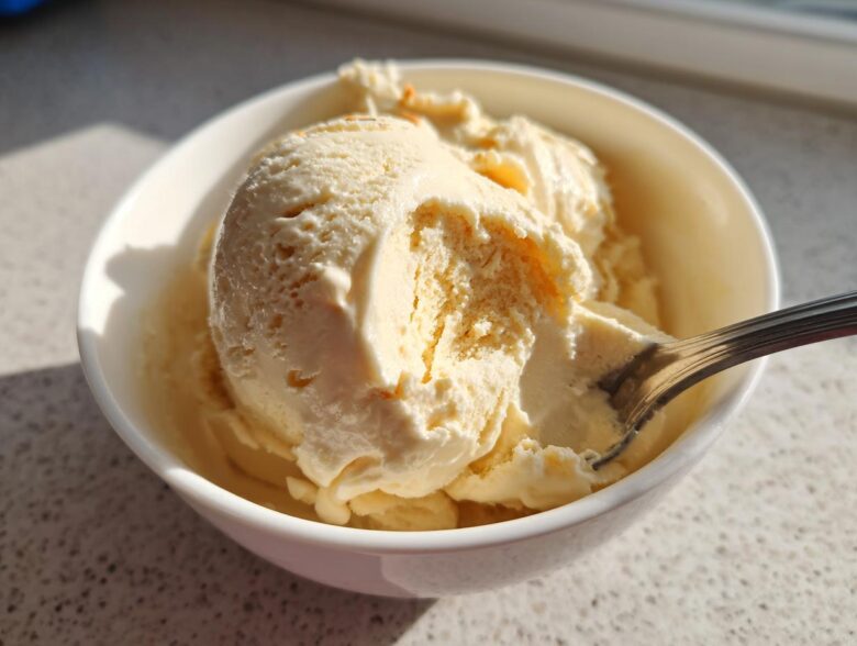 A close-up of creamy, pale yellow banana ice cream scooped into a white bowl with a spoon resting in it.
