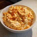 A close-up of creamy, orange-tinted boat dip topped with paprika and herbs in a white serving bowl.