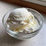 Two scoops of creamy, pale yellow cottage cheese ice cream served in a small, clear glass bowl.