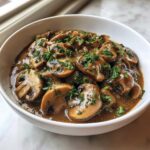 Close-up of a white bowl filled with rich, brown mushroom sauce and sliced mushrooms, garnished with fresh parsley.