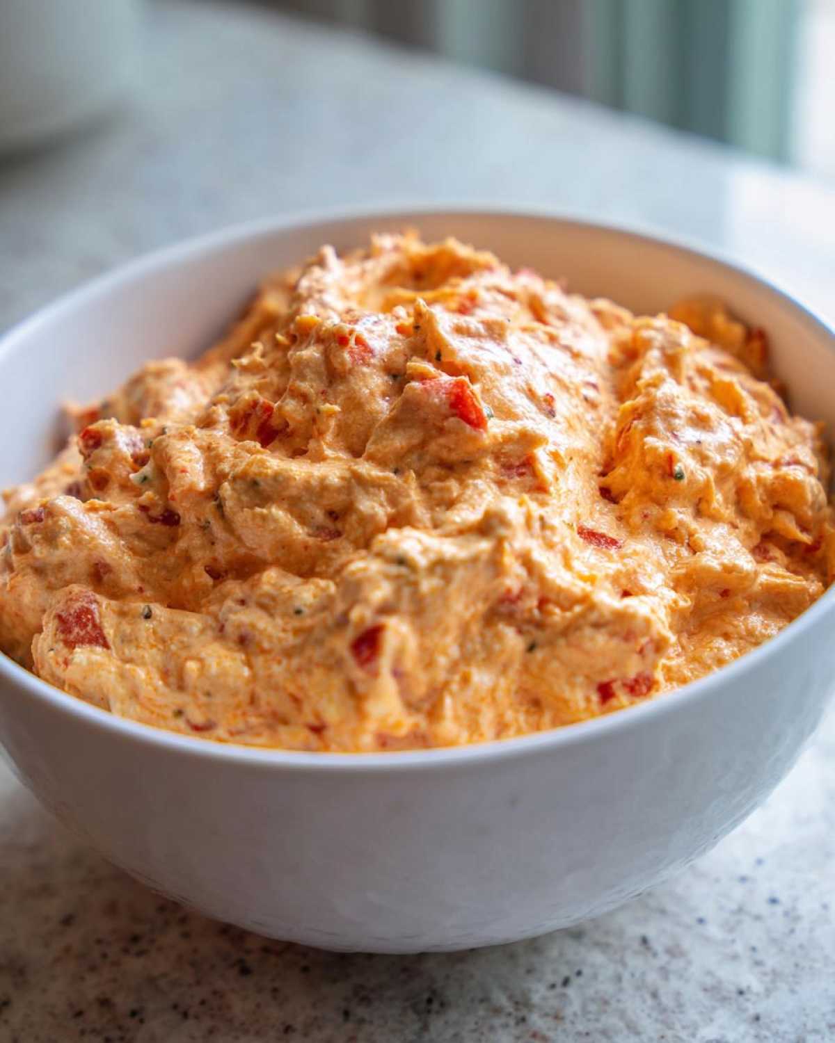 Close-up of a thick, creamy orange boat dip mixed with red pepper pieces, served in a white bowl.
