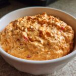 A close-up of creamy, orange-colored boat dip mixed with red pepper flakes and spices, served in a white bowl.