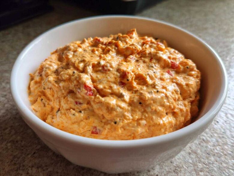 A close-up of creamy, orange-colored boat dip mixed with red pepper flakes and spices, served in a white bowl.