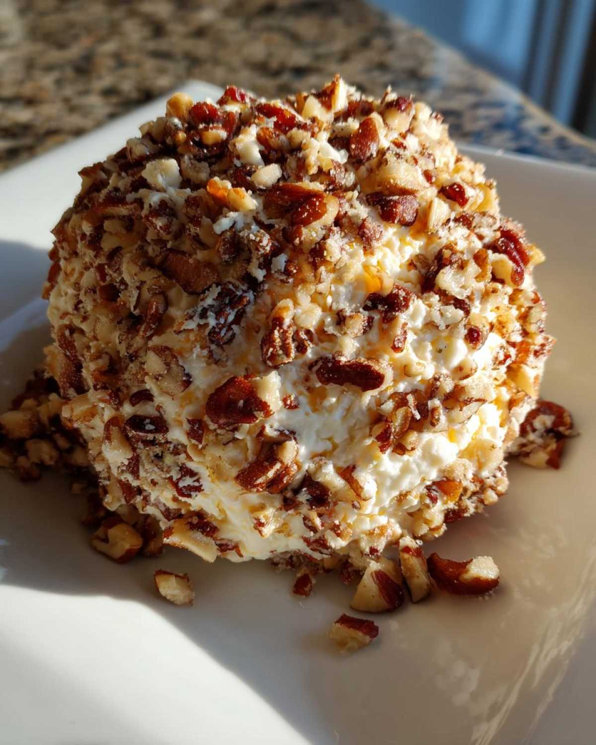 A creamy, round cheese ball recipe coated entirely in chopped pecans, sitting on a white plate.