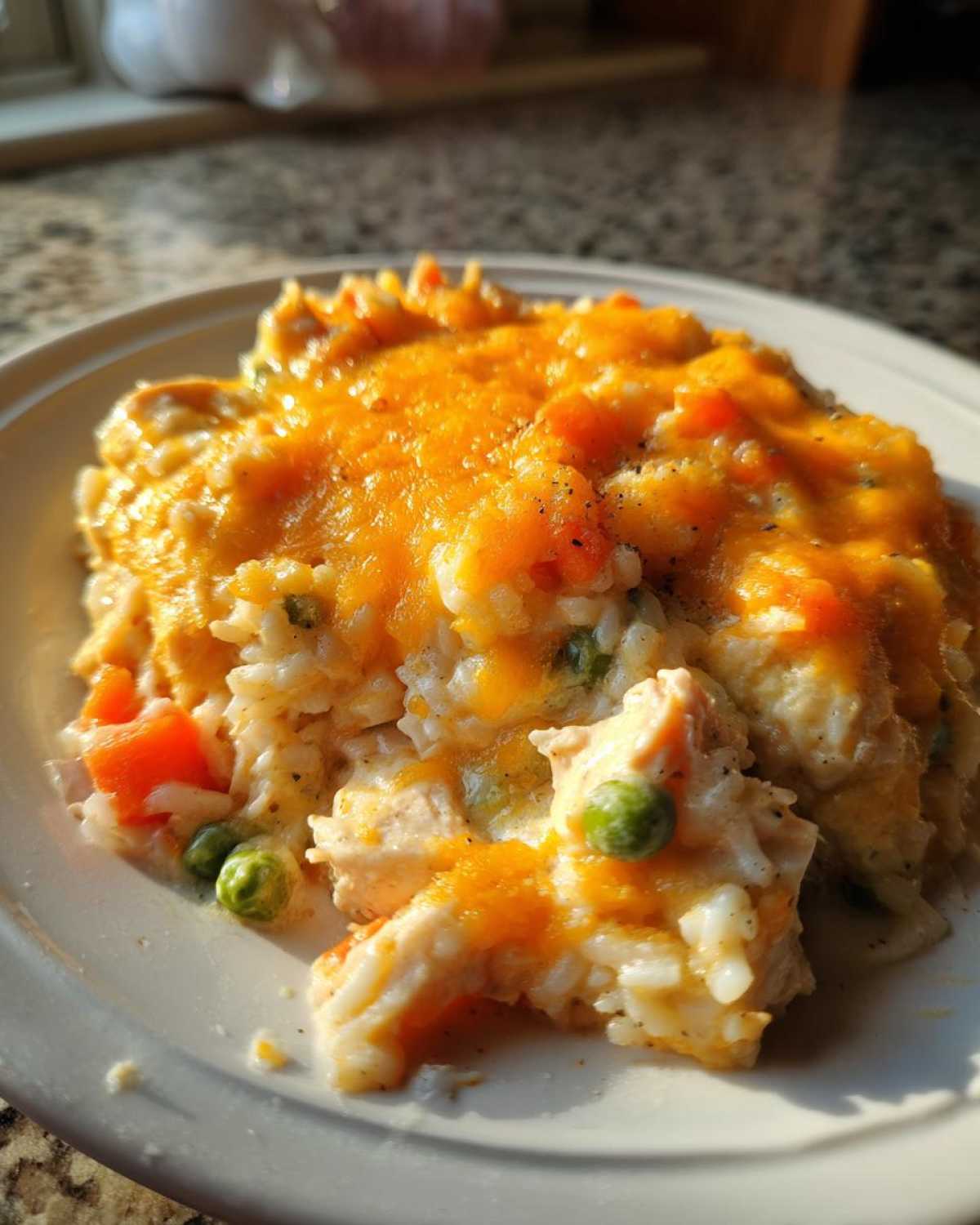 A close-up serving of creamy rice casserole topped with melted cheddar cheese, peas, and carrots.