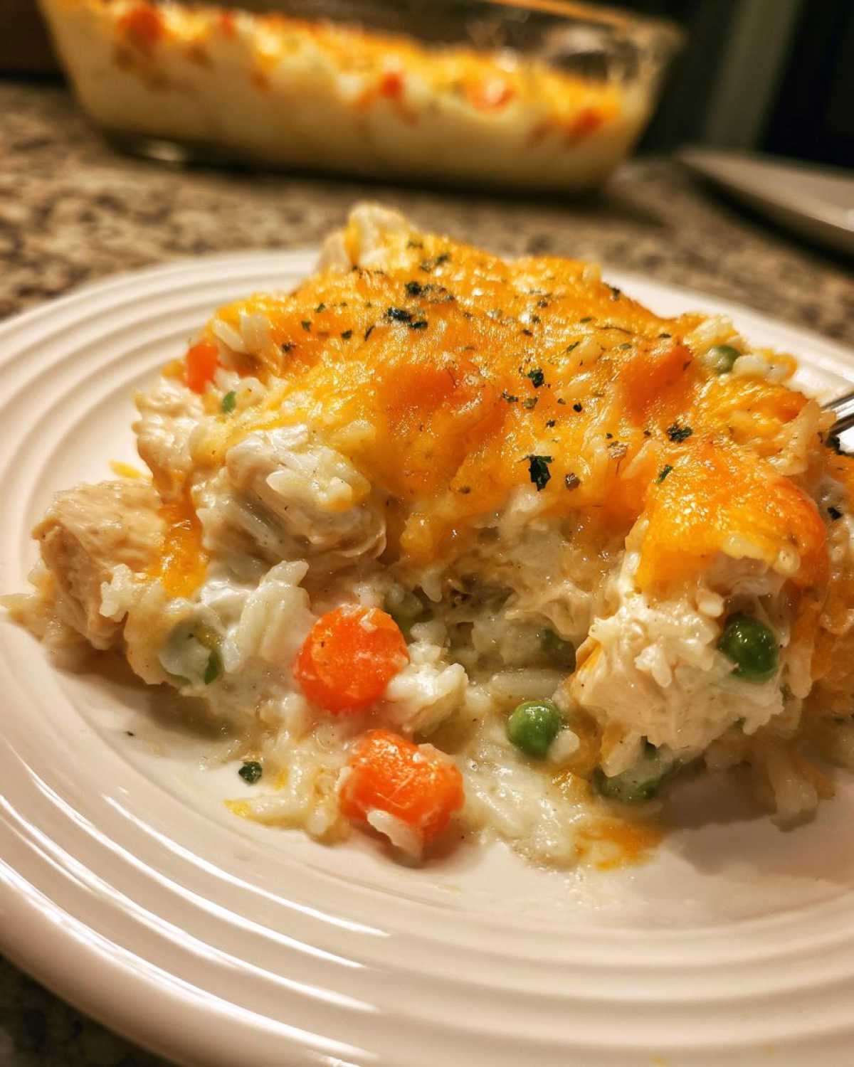 A close-up serving of creamy rice casserole topped with melted cheddar cheese, peas, and carrots on a white plate.