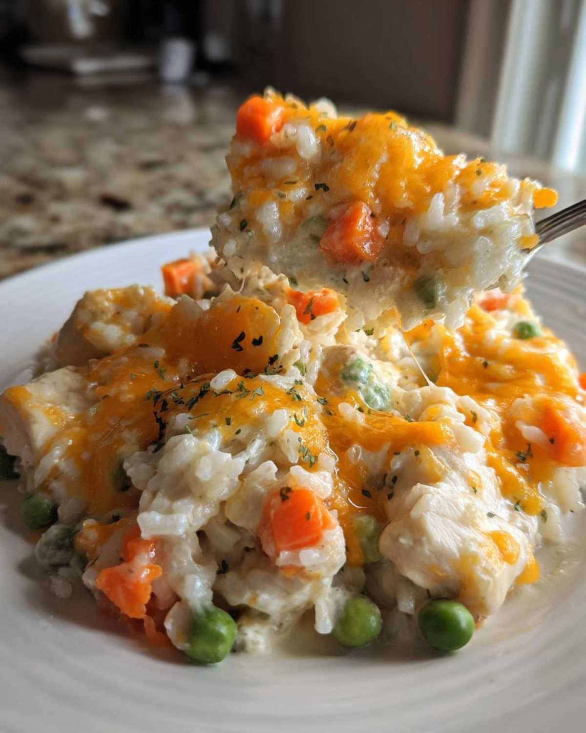 A spoonful of creamy rice casserole with chicken, peas, carrots, and melted cheddar cheese being lifted from a plate.