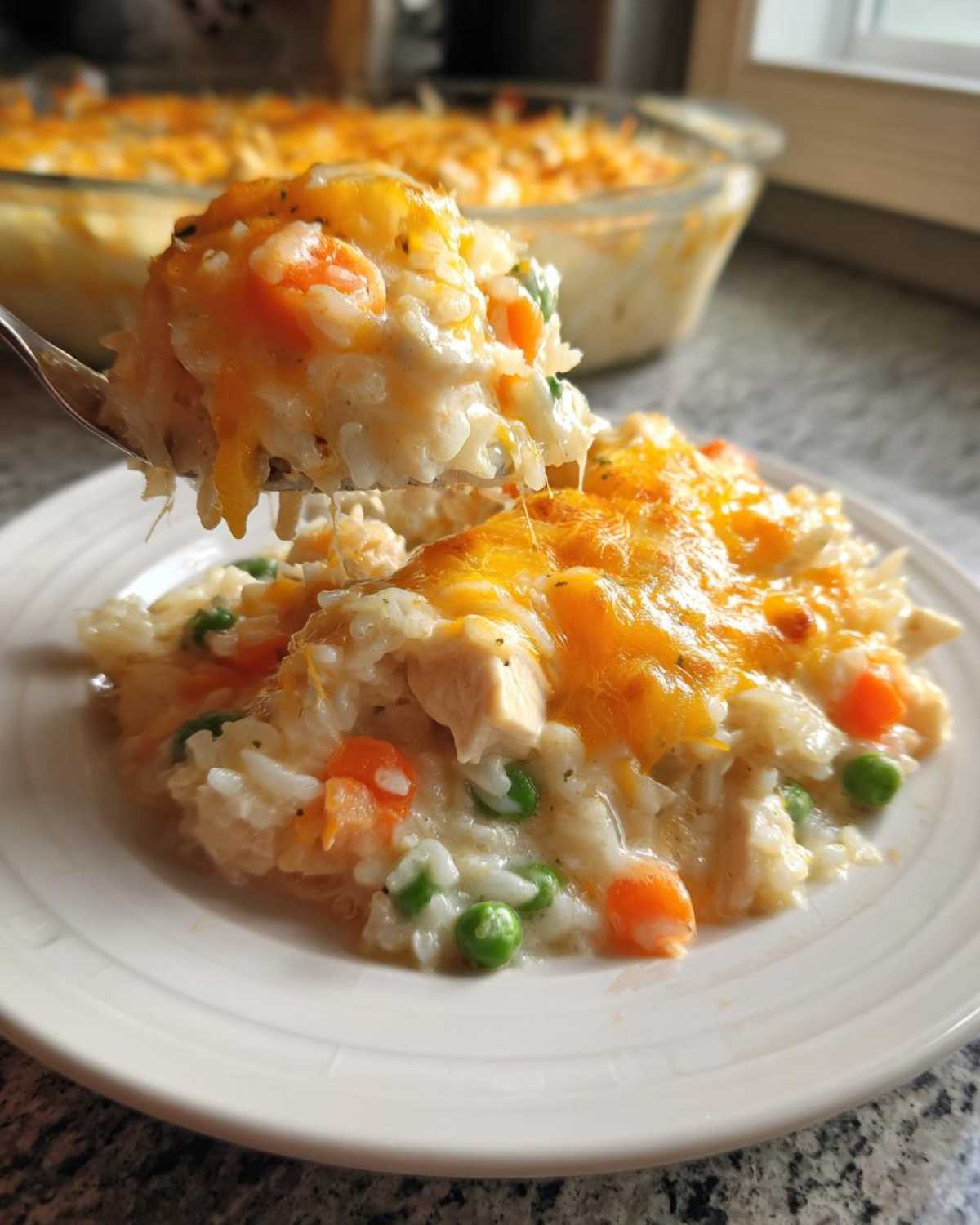 A spoonful of creamy rice casserole with chicken, peas, and carrots, topped with melted cheddar cheese, being lifted from a serving on a plate.