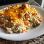 A close-up serving of creamy rice casserole topped with melted cheddar cheese, peas, and carrots on a white plate.