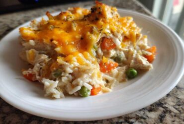 A close-up serving of creamy rice casserole topped with melted cheddar cheese, peas, and carrots on a white plate.